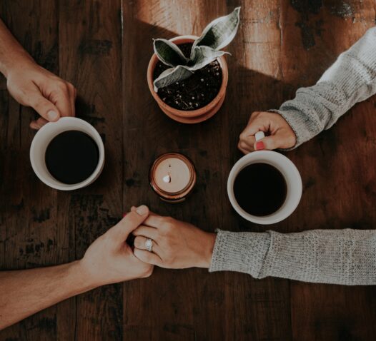 Tre händer runt två kaffekoppar och en krukväxt på ett träbord.